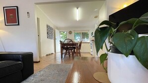 Uma sala de estar moderna com piso de madeira, um sofá de couro preto, uma área de jantar com mesa e cadeiras de madeira, e uma grande planta em vaso.
