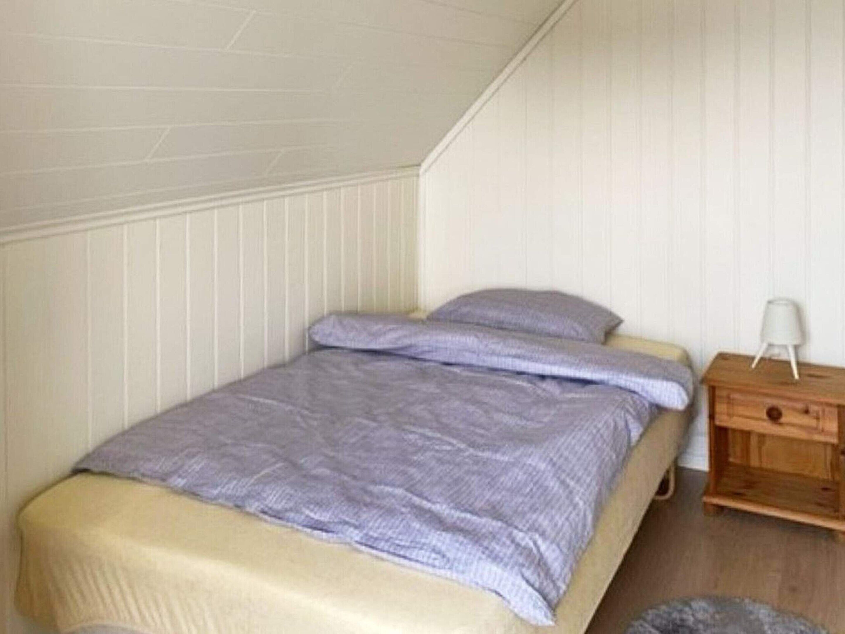A small, single-bed room with a sloped ceiling, white paneled walls, a wooden nightstand, and a bedside lamp.