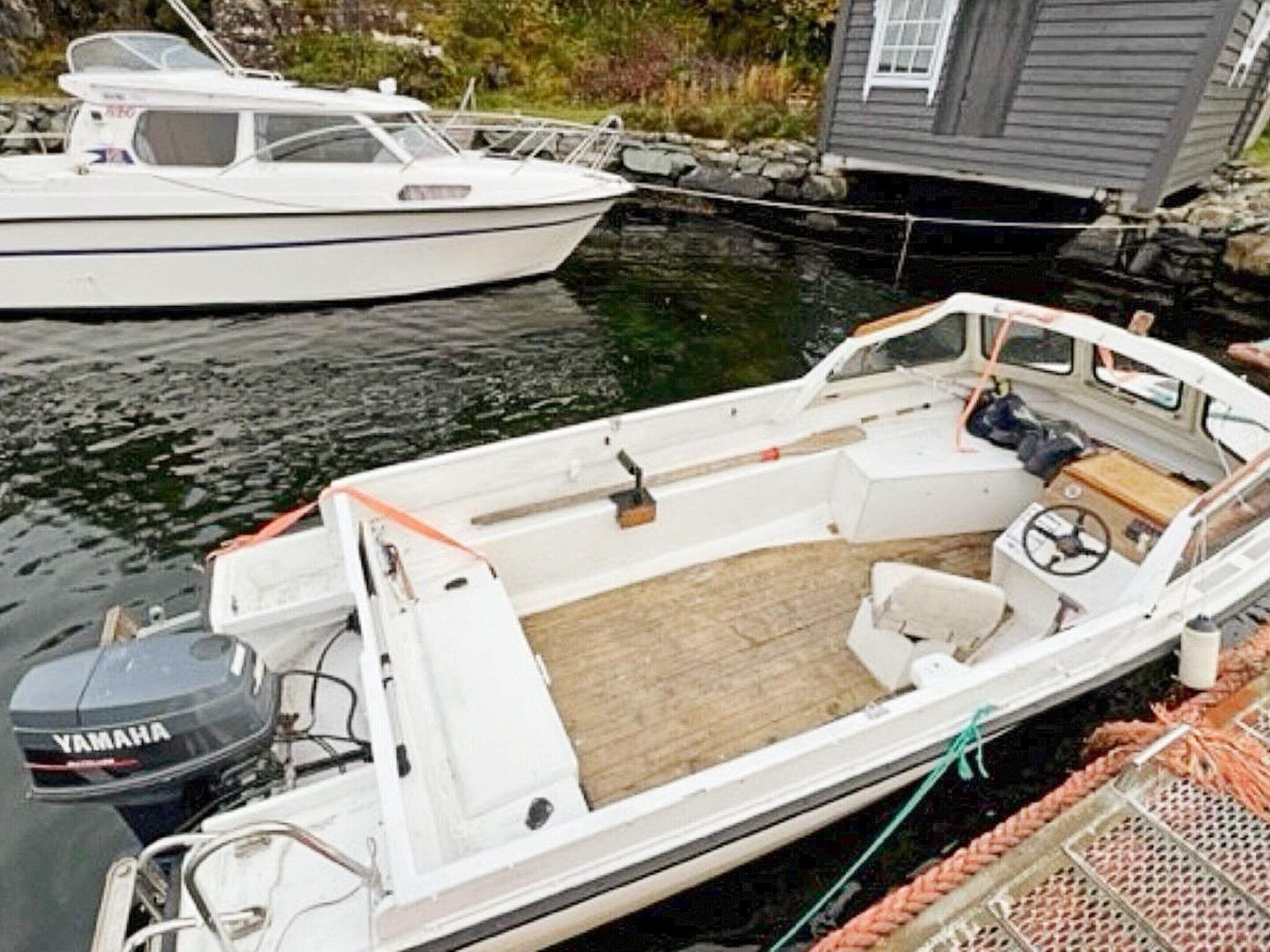 Un petit bateau à moteur blanc était amarré à un quai. Il était équipé d’un moteur hors-bord Yamaha, d’un pont en bois et d’une cabine avec un volant.