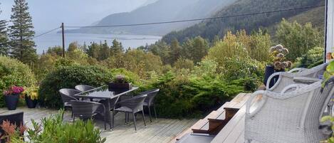 A deck with outdoor furniture overlooking a mountainous landscape and a body of water.