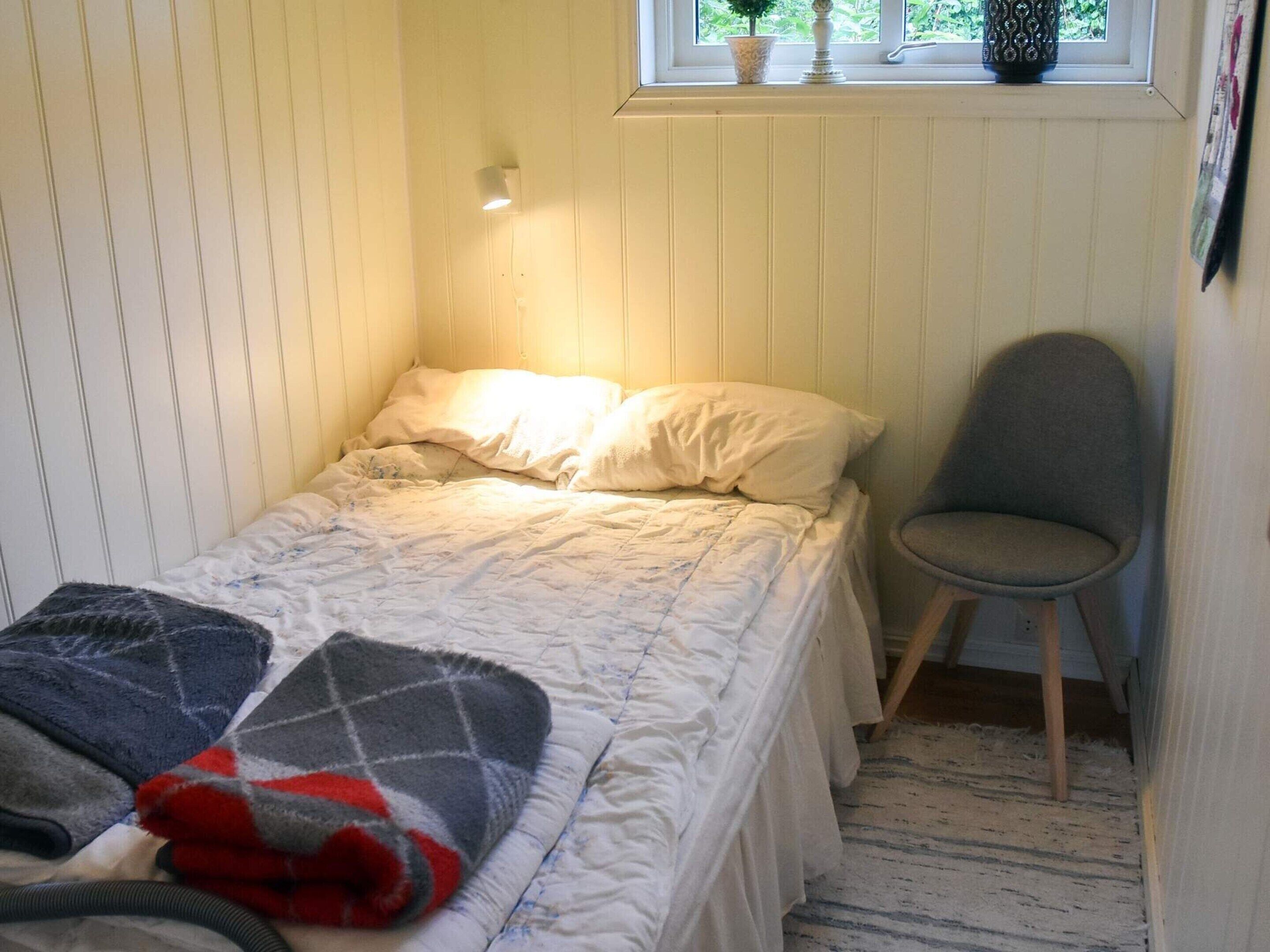 A small, cozy room with a single bed, a chair, a window with a view of greenery, and a wall-mounted light.