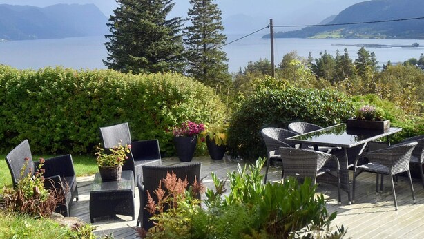An outdoor patio with wicker furniture, potted plants, and a view of a lake and mountains. - 4 Star Holiday Home in Aram (Vanylven)