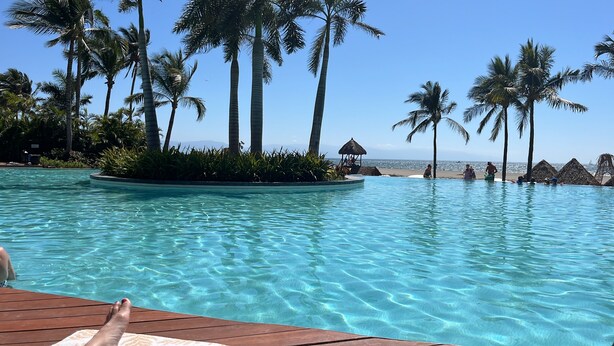 Une piscine aux eaux cristallines, dotée d’une terrasse en bois, entourée de palmiers et offrant une vue sur la plage.