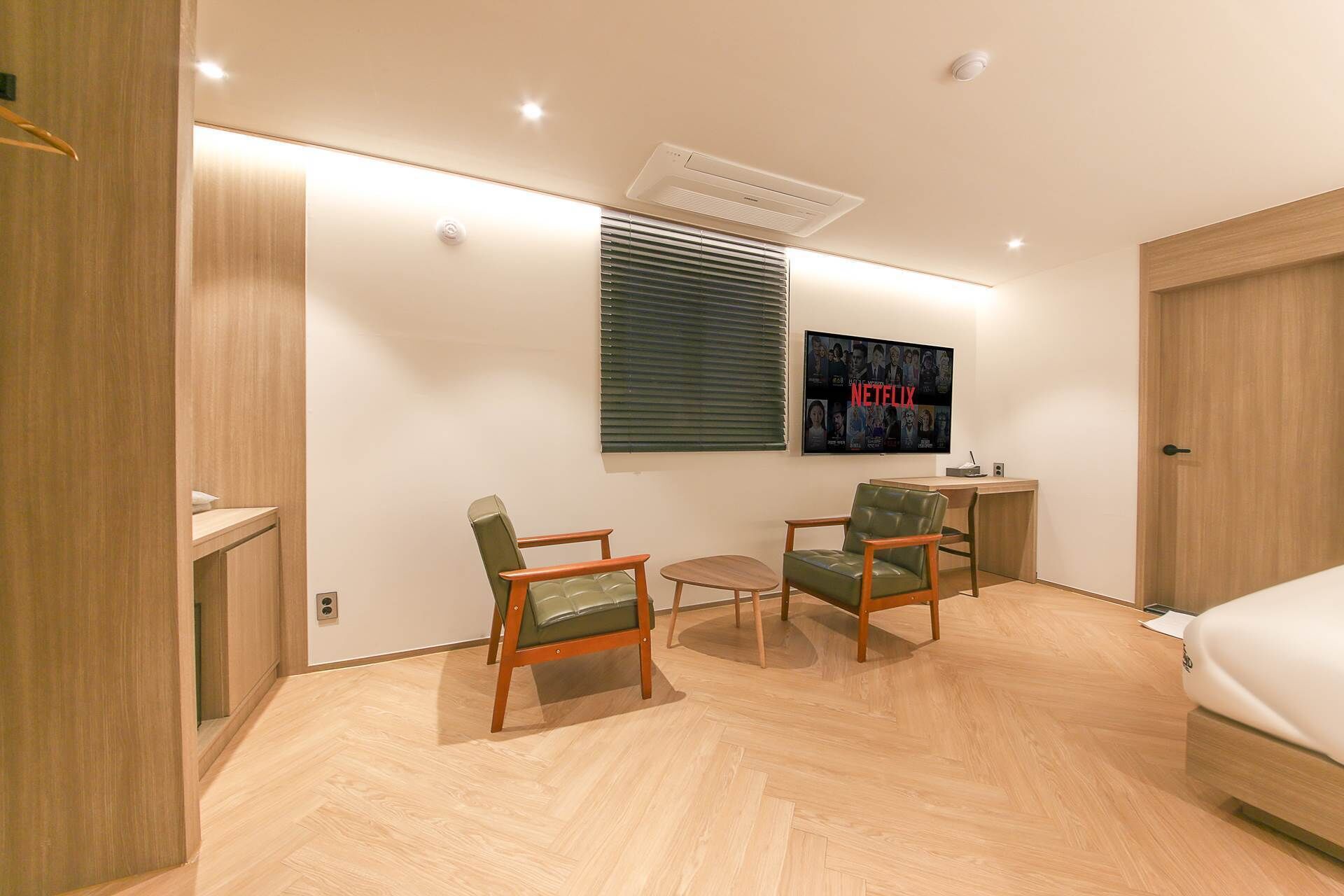 Una habitación de hotel moderna con suelo de madera, dos sillones tapizados en verde, una pequeña mesa redonda, una cama, un escritorio con una computadora y un aire acondicionado de pared, además de un cartel con el logotipo de Netflix.