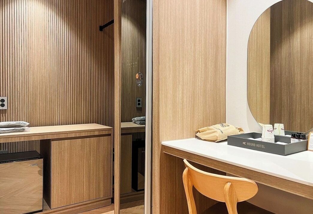 A modern hotel room with a wooden panel wall, a white desk with a mirror, a small wooden chair, and a minibar.