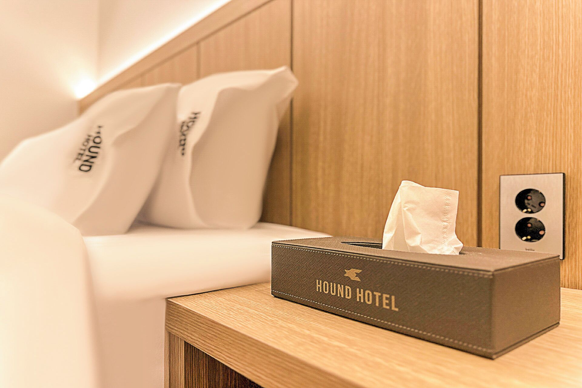 A hotel room with a neatly made bed, a bedside table with a tissue box labeled "HOUND HOTEL," and a wall-mounted light fixture.