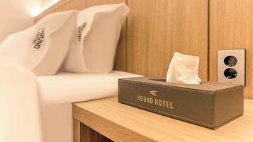 Una habitació d'hotel amb un llit ben arrenjat, una taula de nit amb una caixa de mocadors amb la inscripció "HOUND HOTEL", i una làmpada de paret.