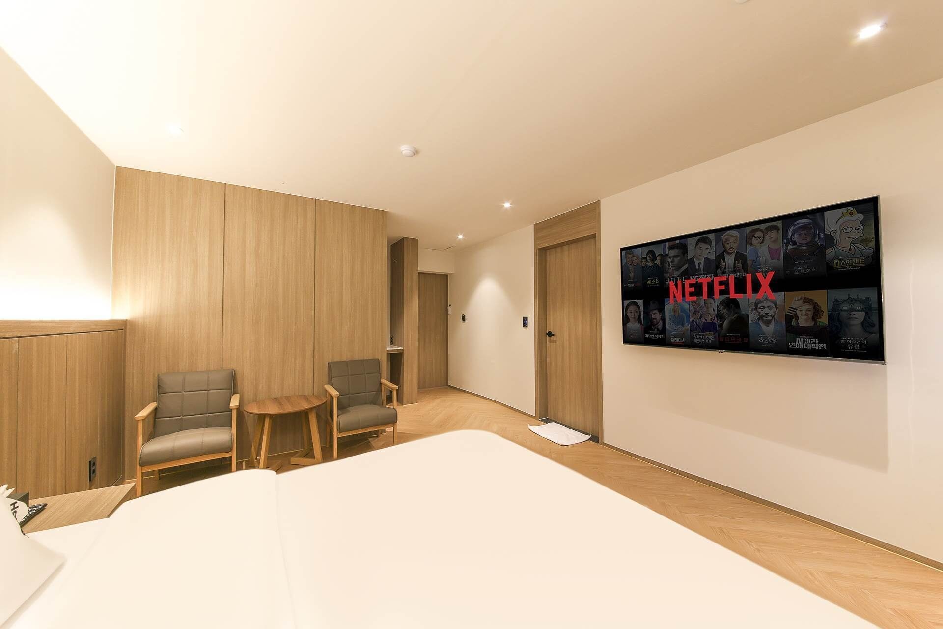 A hotel room with a large wall-mounted TV displaying Netflix content, a wooden panel wall, and two armchairs.