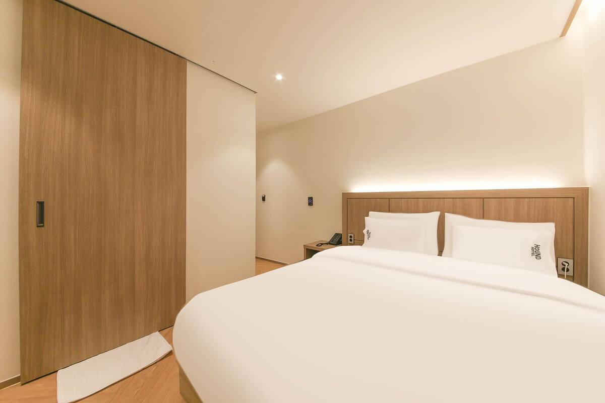 Habitación de hotel con una cama grande, armario de madera, mesita de noche con teléfono y mesita auxiliar.