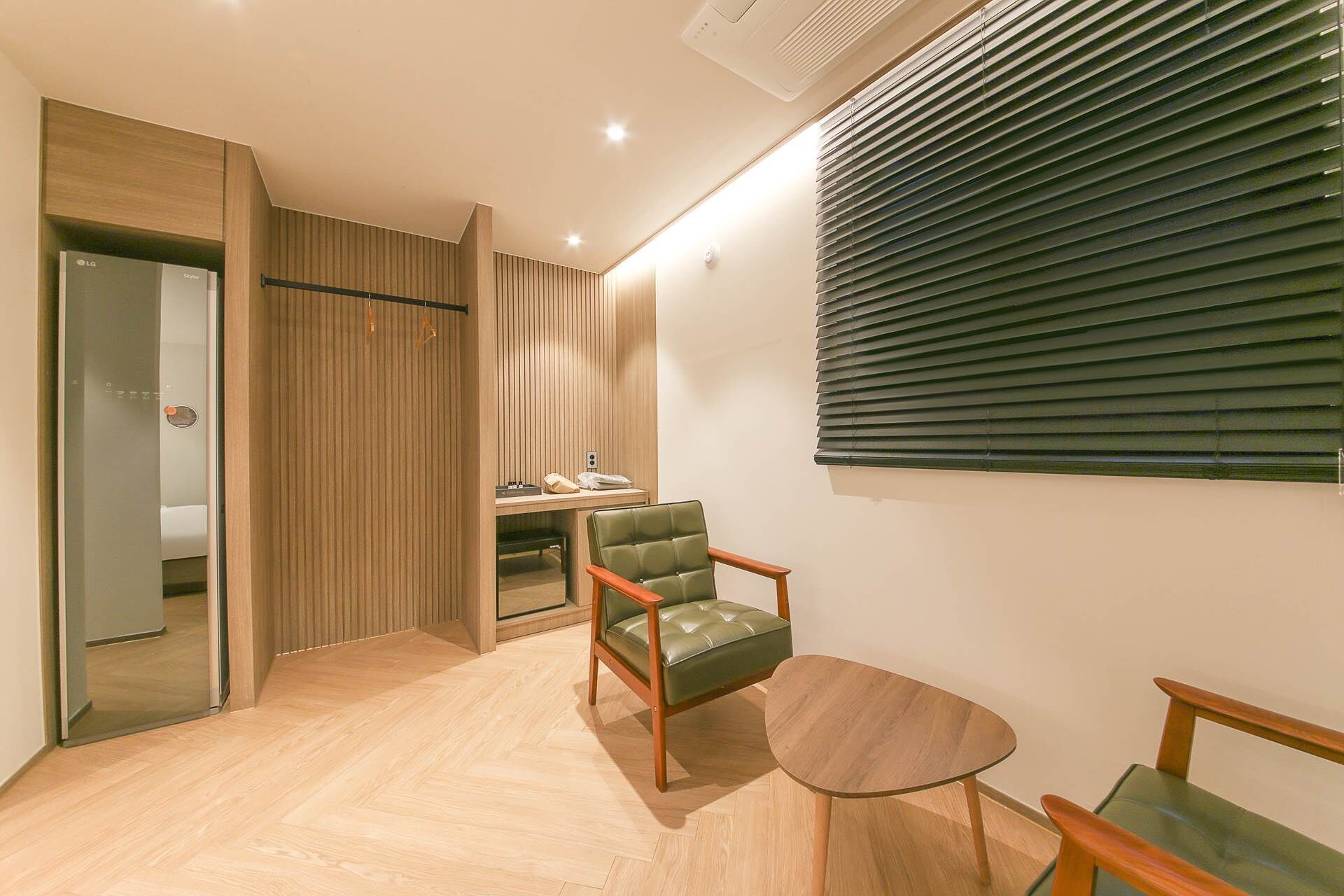 A modern hotel room with a wooden floor, a green leather chair, a small round table, and a desk with a lamp.