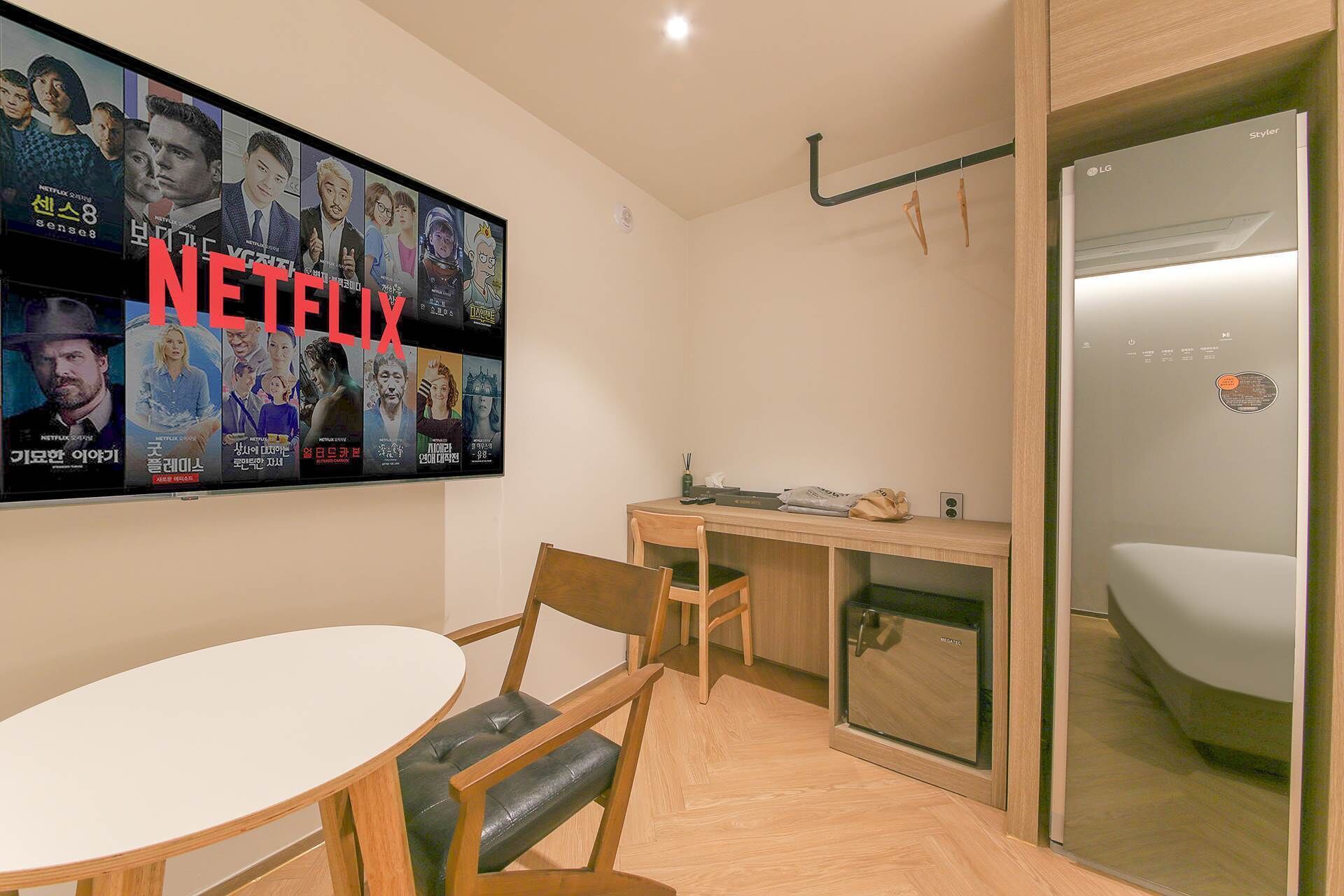 Una habitación de hotel moderna con un televisor grande montado en la pared que muestra contenido de Netflix, una mesa de comedor de madera con sillas, una pequeña kitchenette y un área de ducha con paredes de vidrio.