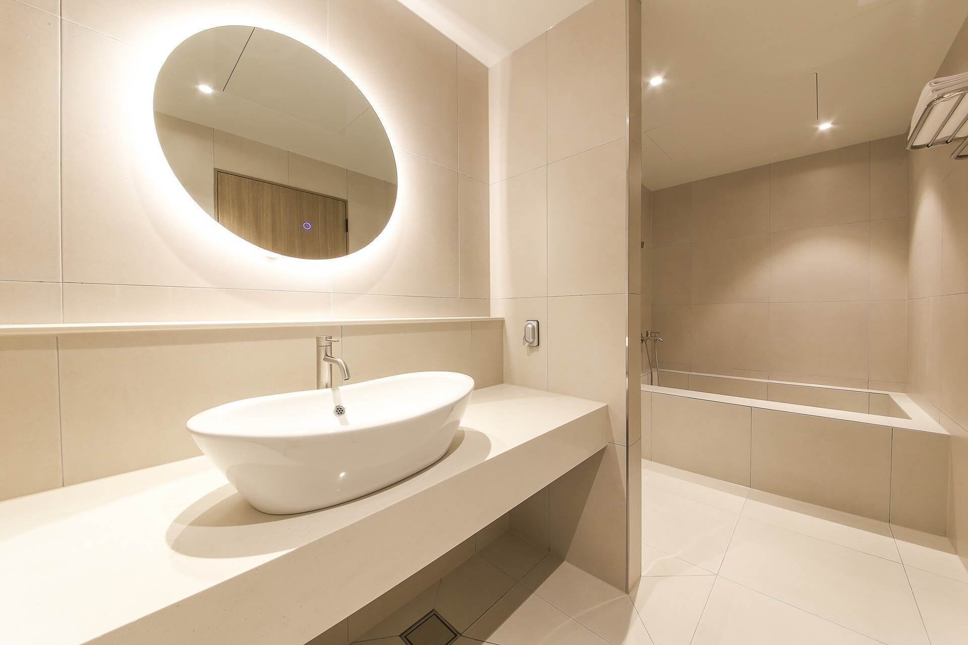 A modern bathroom with a large oval mirror, a white sink, and a glass-enclosed shower area.