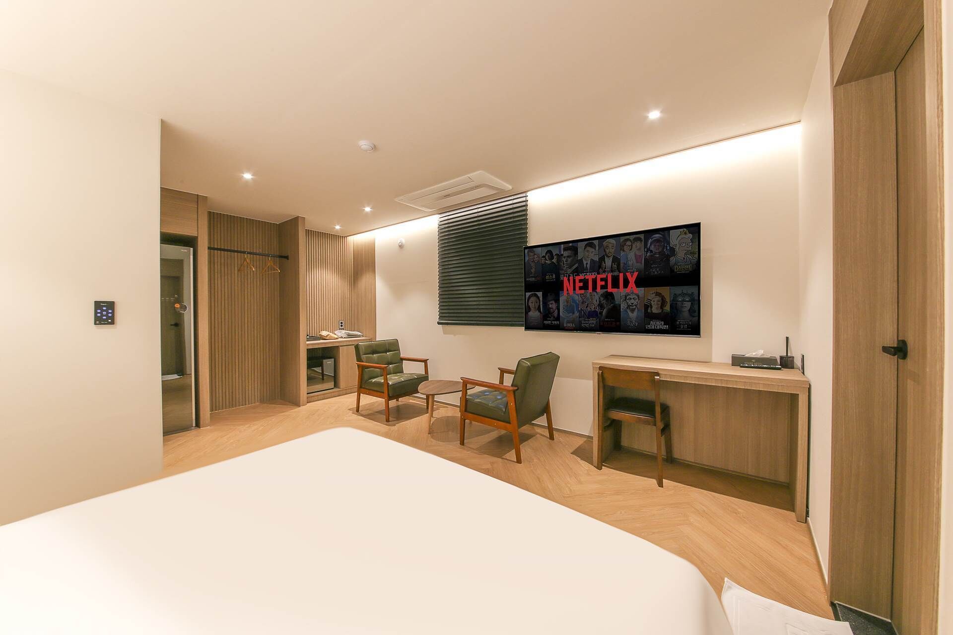 A modern hotel room with a large bed, a desk, two chairs, a TV mounted on the wall, and a wardrobe.