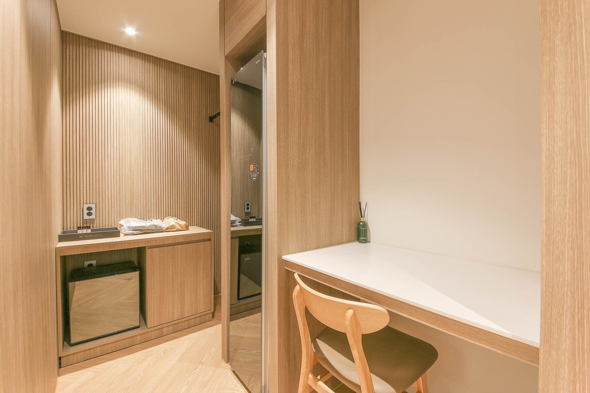 A modern hotel room with a wooden panel wall, a desk with a chair, and a small table with a bottle of water and a tissue box.
