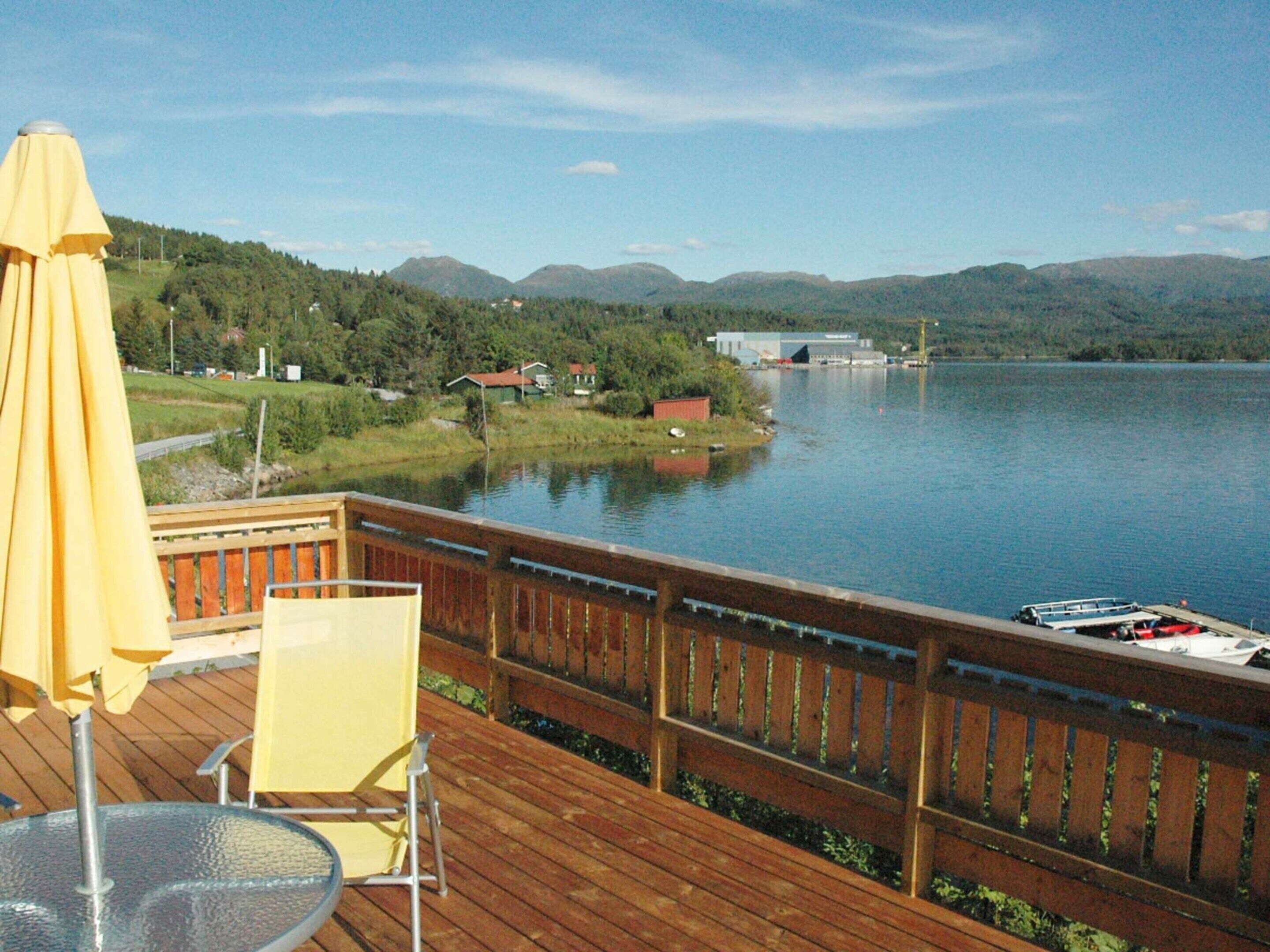A wooden deck with a table and chairs, overlooking a calm lake with mountains in the background.