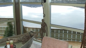 A room with large windows overlooking a lake and mountains. There is a wooden chair and a small table with candles and a decorative item.
