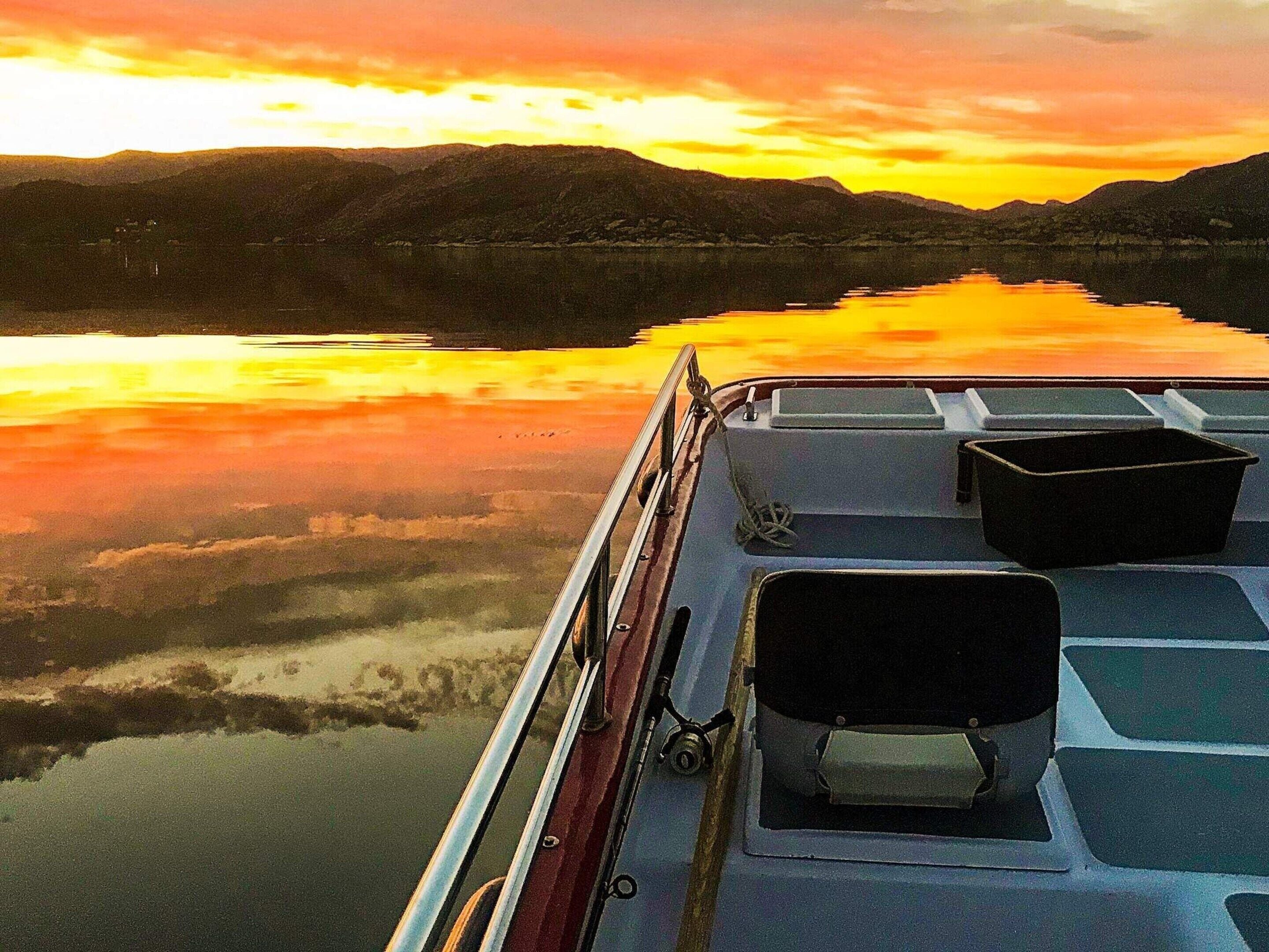Een boot met een metalen reling en twee zwarte zitplaatsen aan de boeg, gericht op een kalm meer met een zonsondergang die in het water weerspiegelt, omgeven door heuvels.