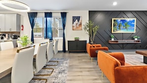 Un comedor moderno con una mesa de madera y sillas blancas, un mueble de almacenamiento oscuro y un amplio ventanal con cortinas azules.