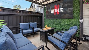 Un patio al aire libre con una chimenea, asientos azules y una gran pantalla de Netflix.