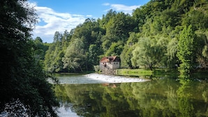 Ein rustikales Steinhaus mit rotem Dach liegt an einem ruhigen Fluss, umgeben von dichtem Grün und Bäumen.