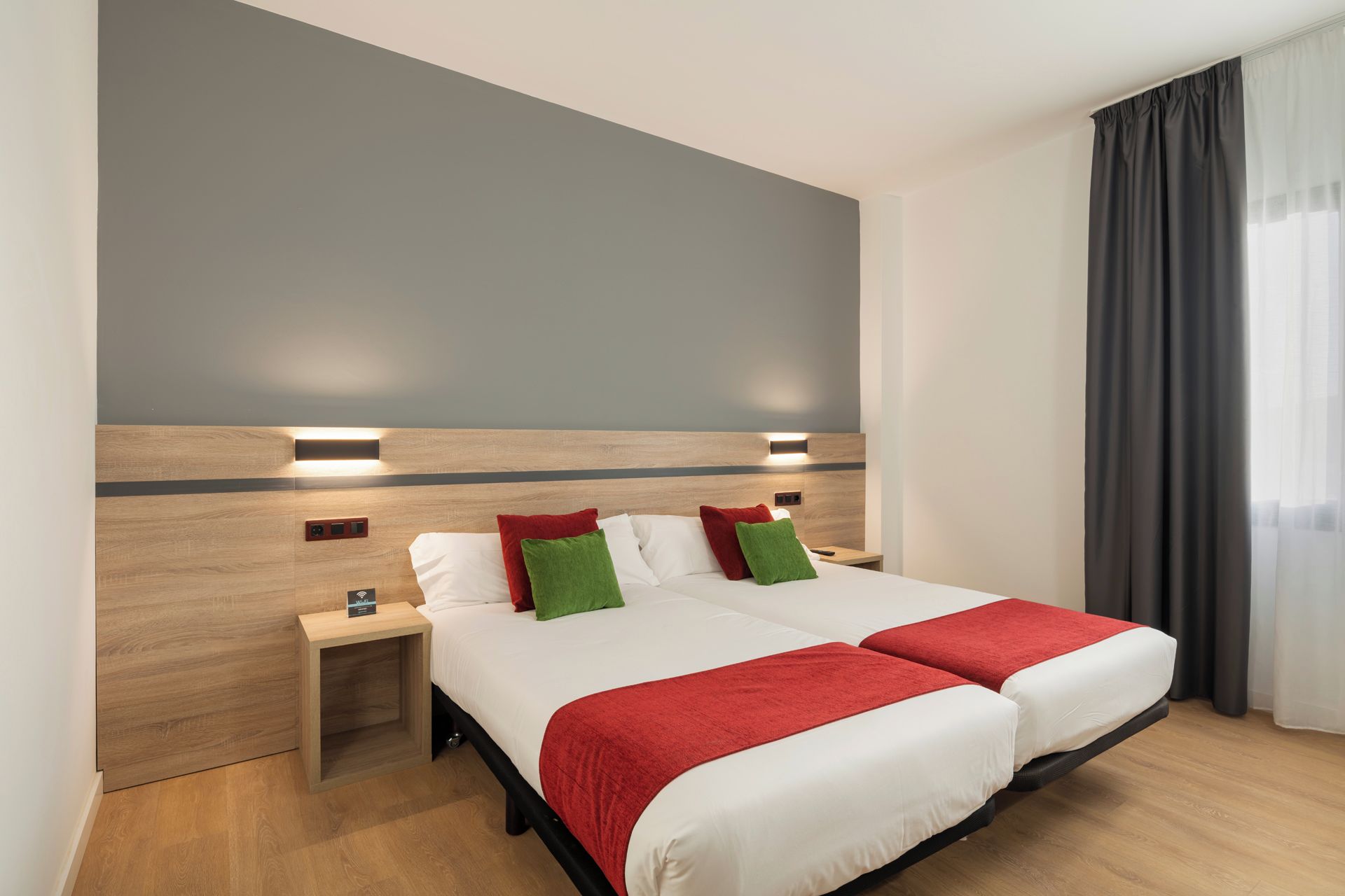 A double bed with two red throw blankets, a nightstand with a lamp, and a grey wall with two wall-mounted lights.