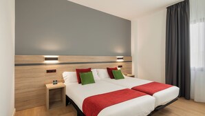 Un lit double avec deux plaids rouges, une table de chevet avec une lampe et un mur gris orné de deux appliques murales.