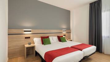 A double bed with two red throw blankets, a nightstand with a lamp, and a grey wall with two wall-mounted lights.