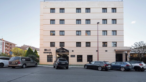A multi-story hotel with a flat facade, a central entrance, and a parking area with several cars. - Alda Malvasia (Haro)