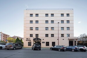 A multi-story hotel with a flat facade, a central entrance, and a parking area with several cars. - Alda Malvasia (Haro)