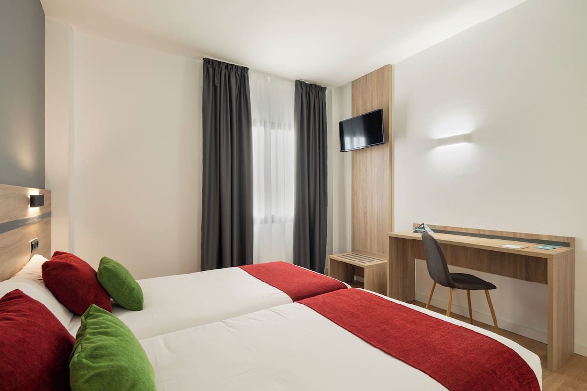 A hotel room with two beds, a desk, a chair, a TV, and a window with curtains.