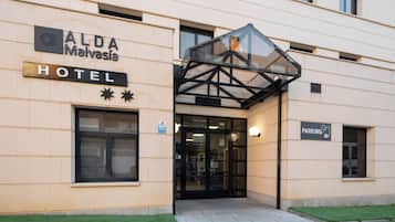 The exterior of ALDA Malasia Hotel with a glass entrance canopy, a sign indicating a parking area, and a small grassy area in front.