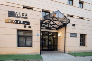 ALDA Malasia Hotel, set med et glas tag over indgangen, et skilt der angiver parkeringsplads, og en lille grøn græsplæne foran.