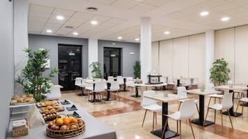 Uma sala de jantar moderna com várias mesas e cadeiras, um buffet com diversos alimentos, e plantas em vasos.