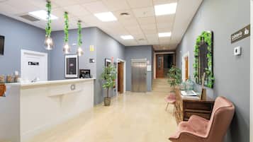 Uma área de recepção com balcão branco, placa indicando sala privativa, mesa de recepção, escada e um relógio fixado na parede.