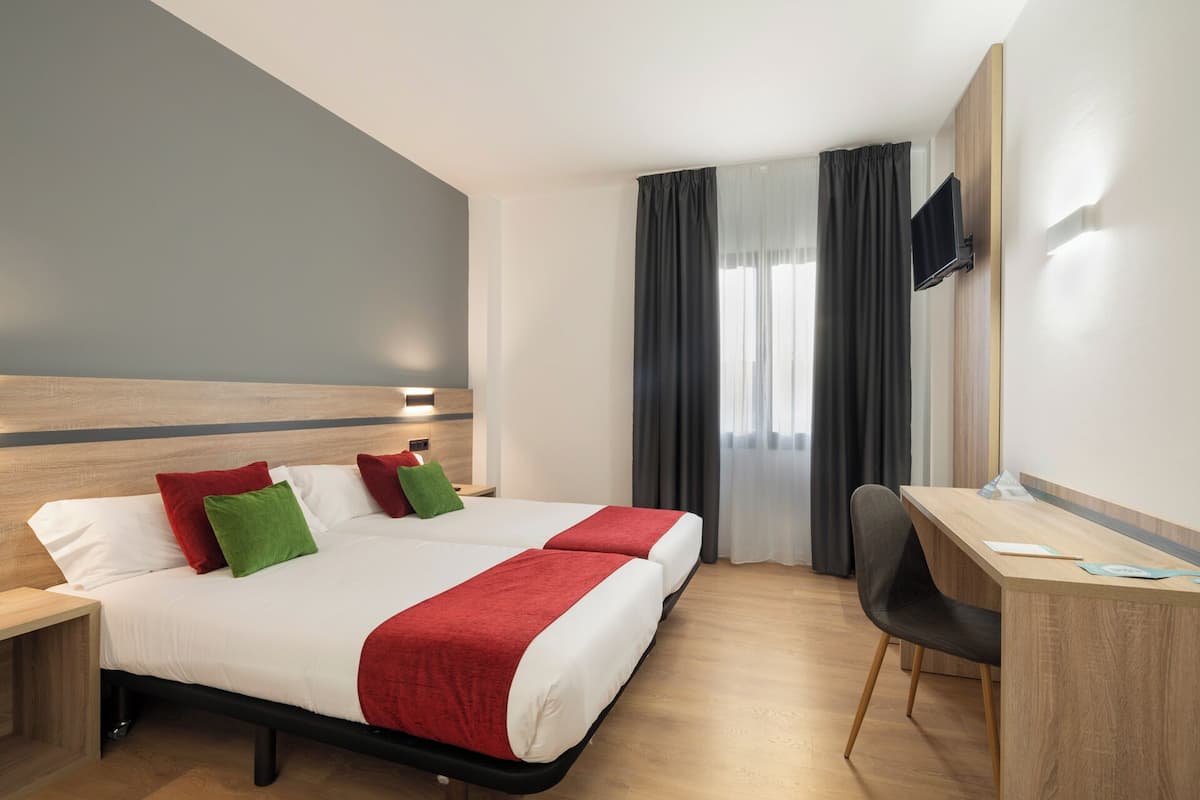 A hotel room with a bed, two bedside tables, a desk, a chair, and a TV mounted on the wall.