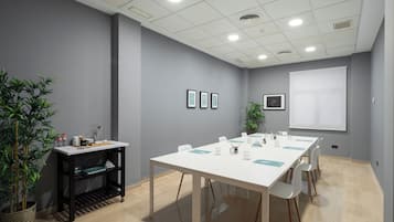 Uma sala de conferências com uma mesa branca, cadeiras, tela de projetor, quadros nas paredes e uma planta.