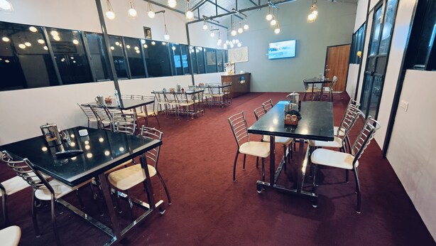 A spacious dining area with multiple tables and chairs, overhead lighting, and a television screen on the wall. - Triyuginarayan Maangal Resort  (Ukhimath)