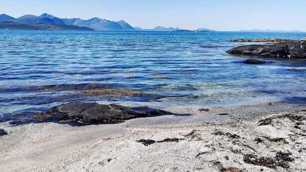 A coastal landscape with clear blue water, rocky shore, and distant mountains. - 6 Person Holiday Home in Borkenes (Kvæfjord)