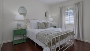 Une chambre à coucher comprenant un lit, deux tables de chevet, un miroir, une fenêtre avec des rideaux et un ventilateur de plafond.