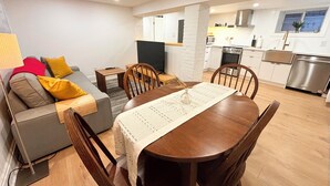 A modern living room with a grey sofa, wooden dining table, and chairs. A kitchen with white cabinets and stainless steel appliances is visible in the background.