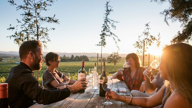Un grupo de personas en una mesa de madera al aire libre, con botellas y copas de vino, rodeado de árboles y un cielo despejado.