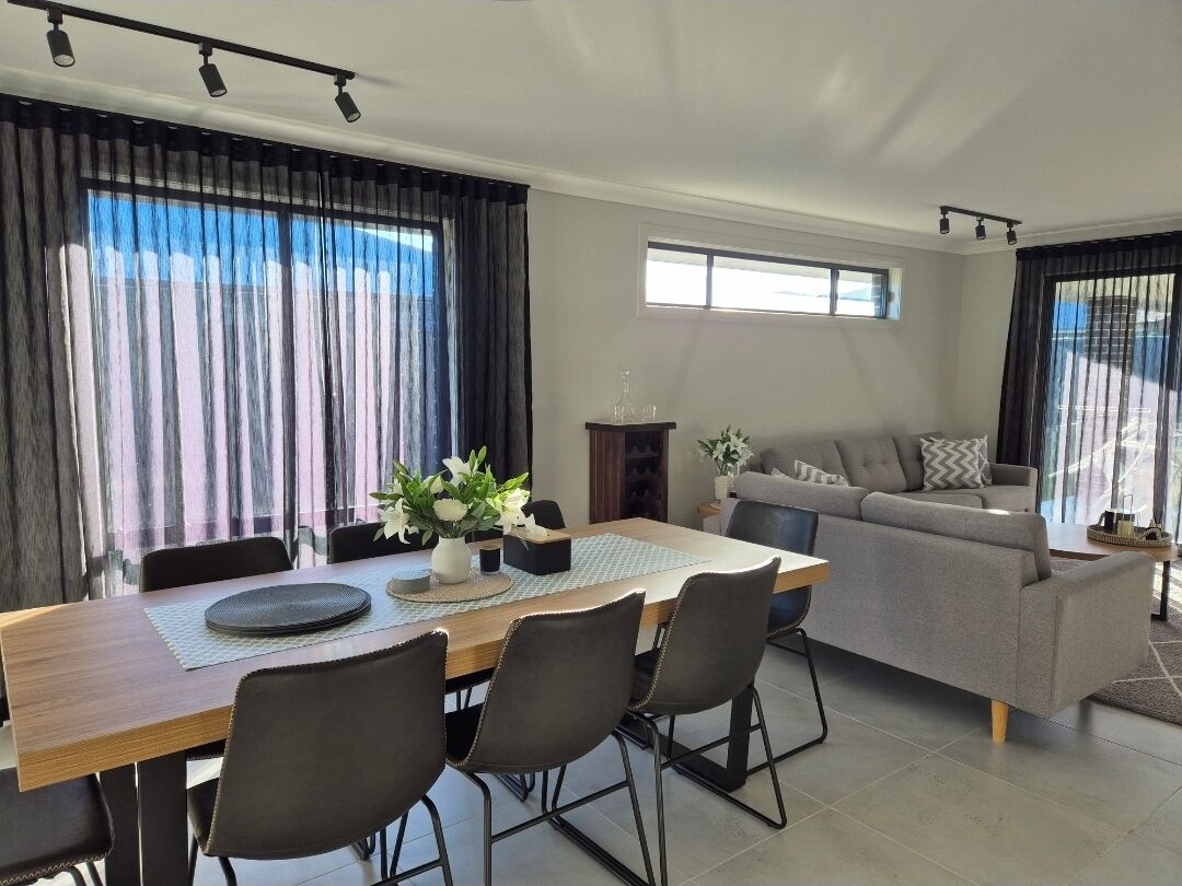 A modern living room with a dining area, featuring a sofa, a dining table with chairs, and large windows with curtains.