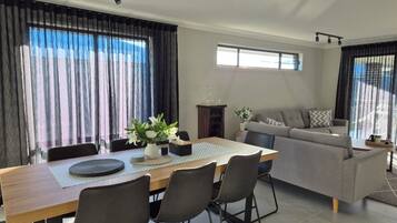 A modern living room with a dining area, featuring a sofa, a dining table with chairs, and large windows with curtains.