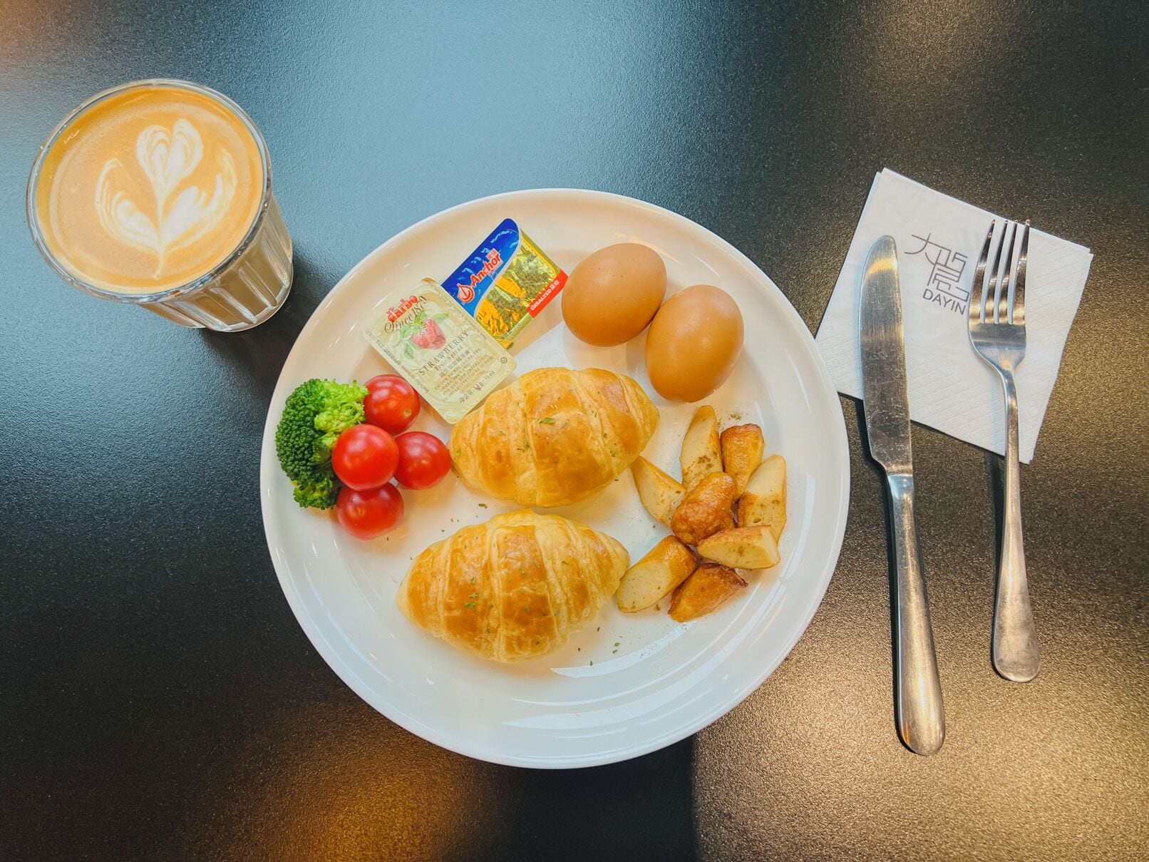 A plate with croissants, eggs, tomatoes, broccoli, and a drink with latte art.