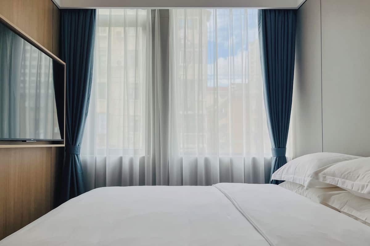 A hotel room with a bed, a TV mounted on the wall, and a window with sheer curtains.