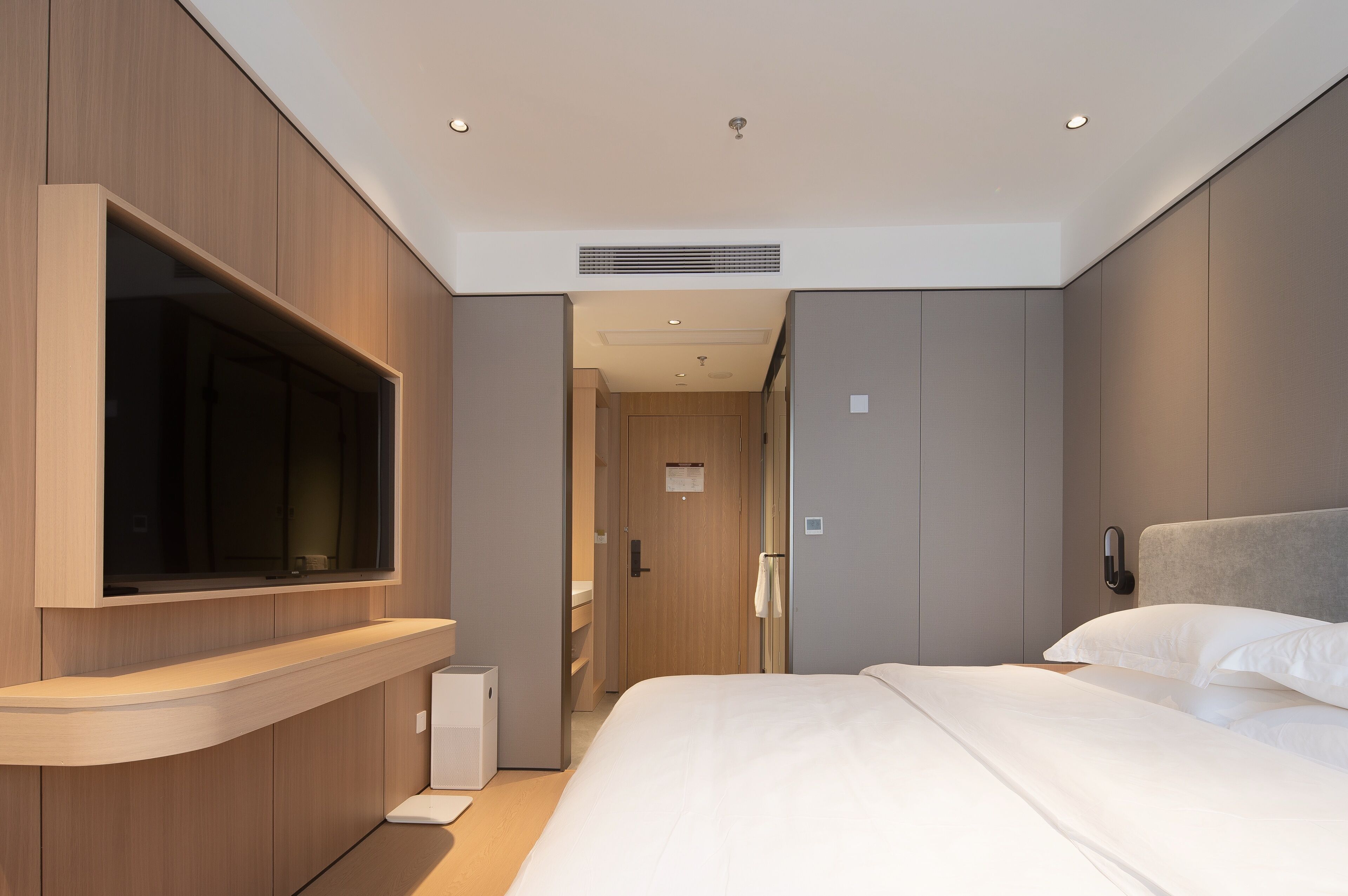 Un cuarto de hotel moderno con una televisión de pantalla plana grande montada en la pared, una cama con sábanas blancas y una mesita de noche.