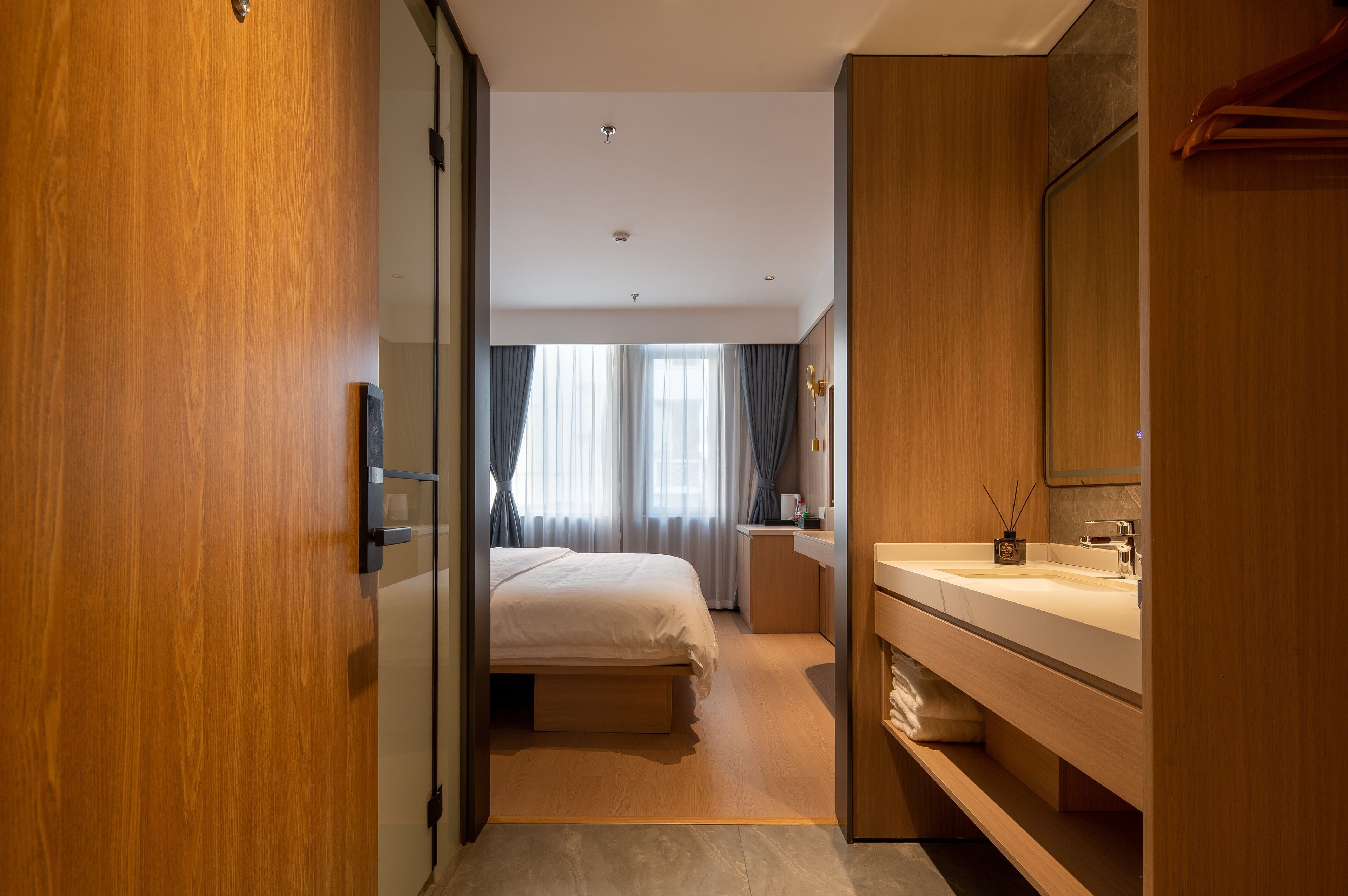 A modern hotel room with a wooden door, a bed, a bathroom with a sink and mirror, and a view of the street through the window.