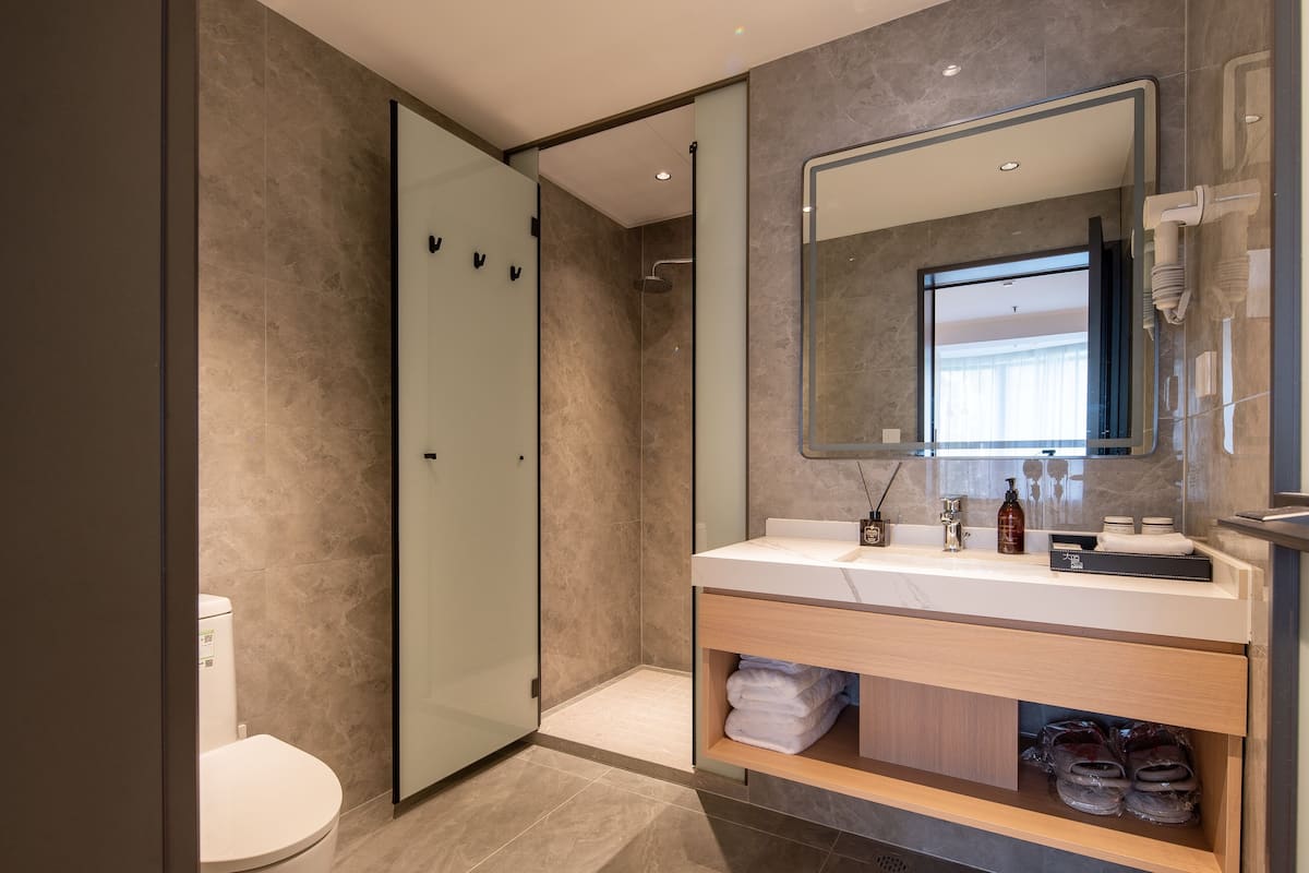 A modern bathroom with a glass shower enclosure, a white toilet, a vanity with a mirror, and a shelf with towels and toiletries.