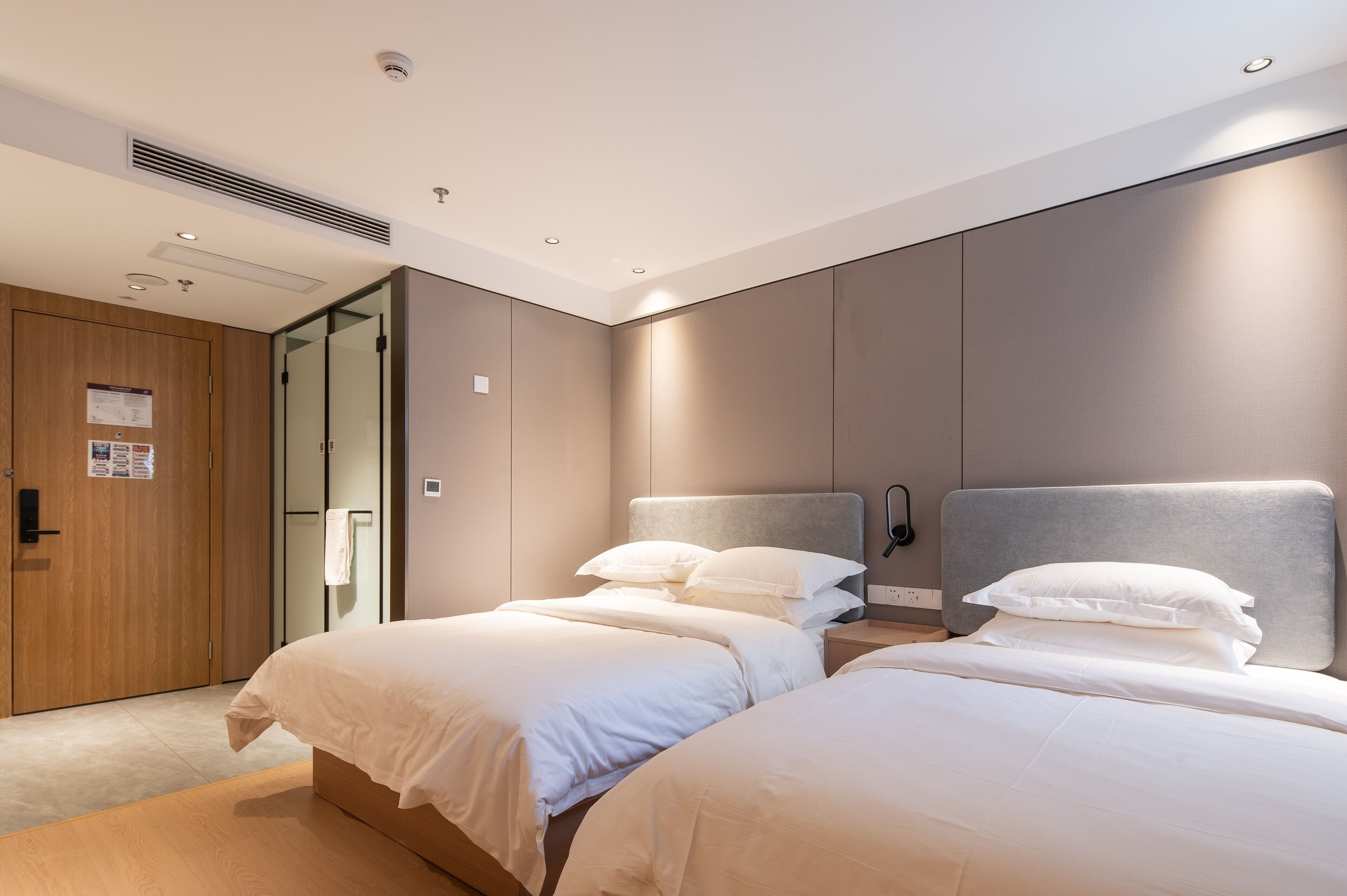 A modern hotel room with two beds, a wooden wardrobe, a glass door, and a wall-mounted light fixture.