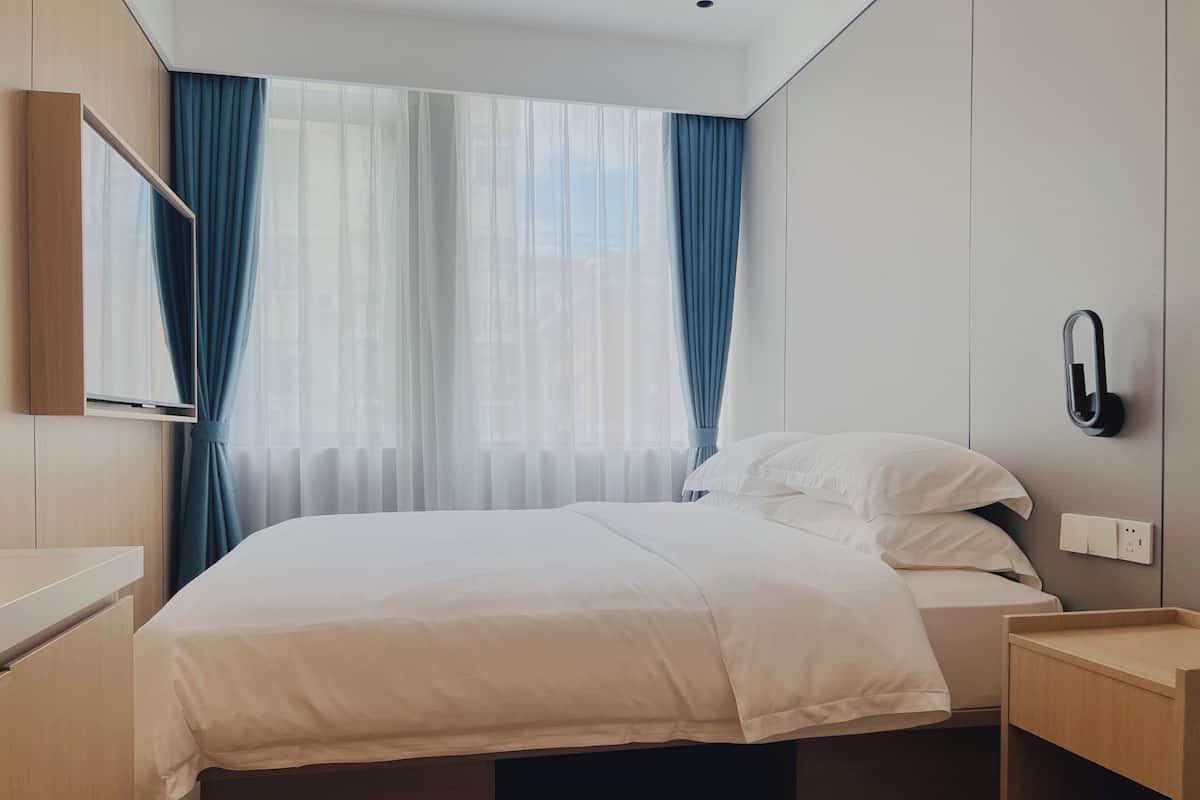 A modern hotel room with a bed, a bedside table, a TV mounted on the wall, and a window with curtains.